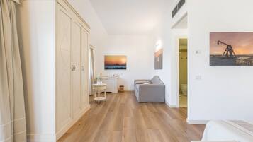 A spacious room with a wooden floor, a grey sofa, a small table with a bottle, and a painting of a sunset on the wall.