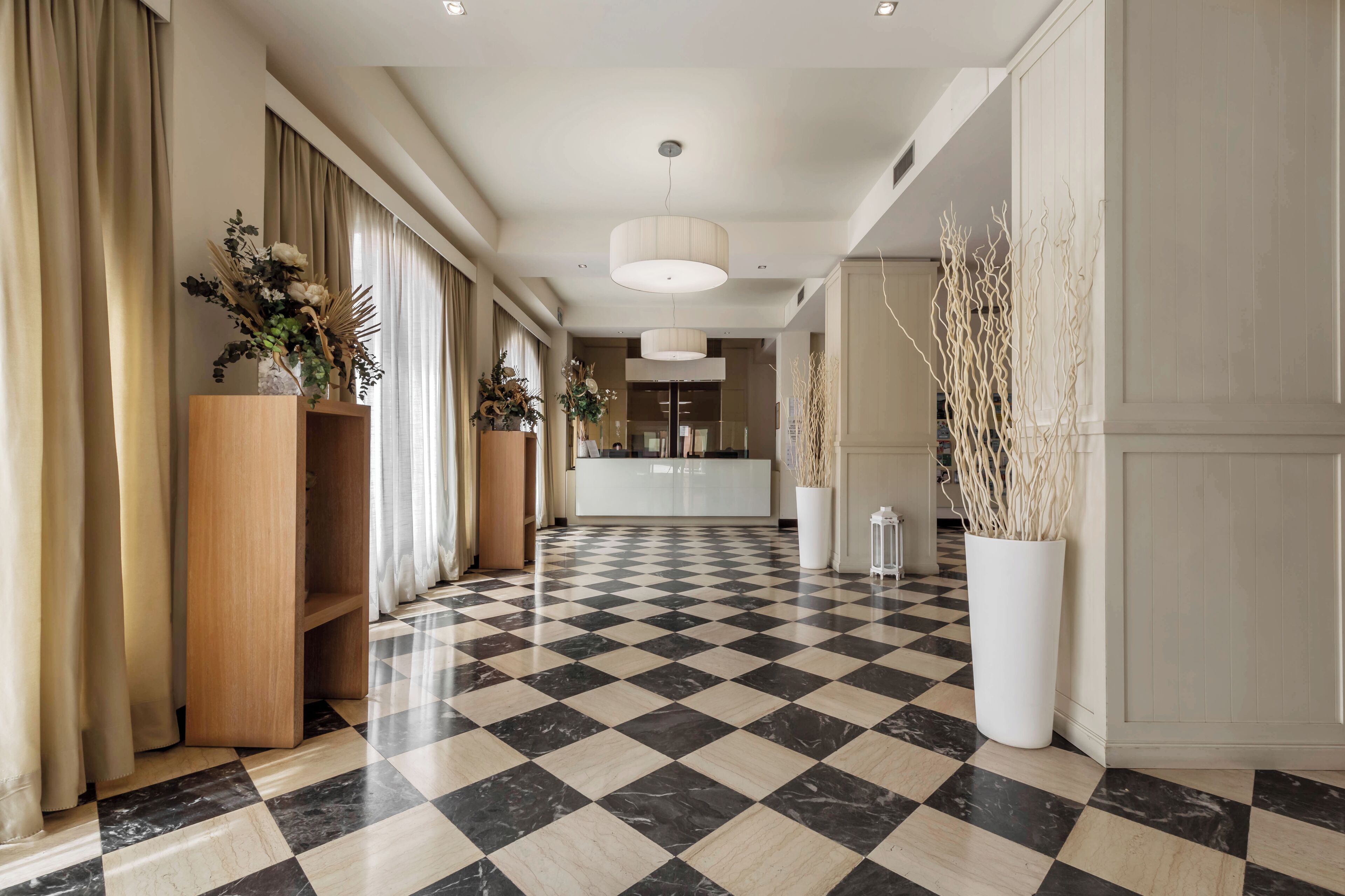 A hotel lobby with a checkered floor, wooden reception desk, and potted plants.