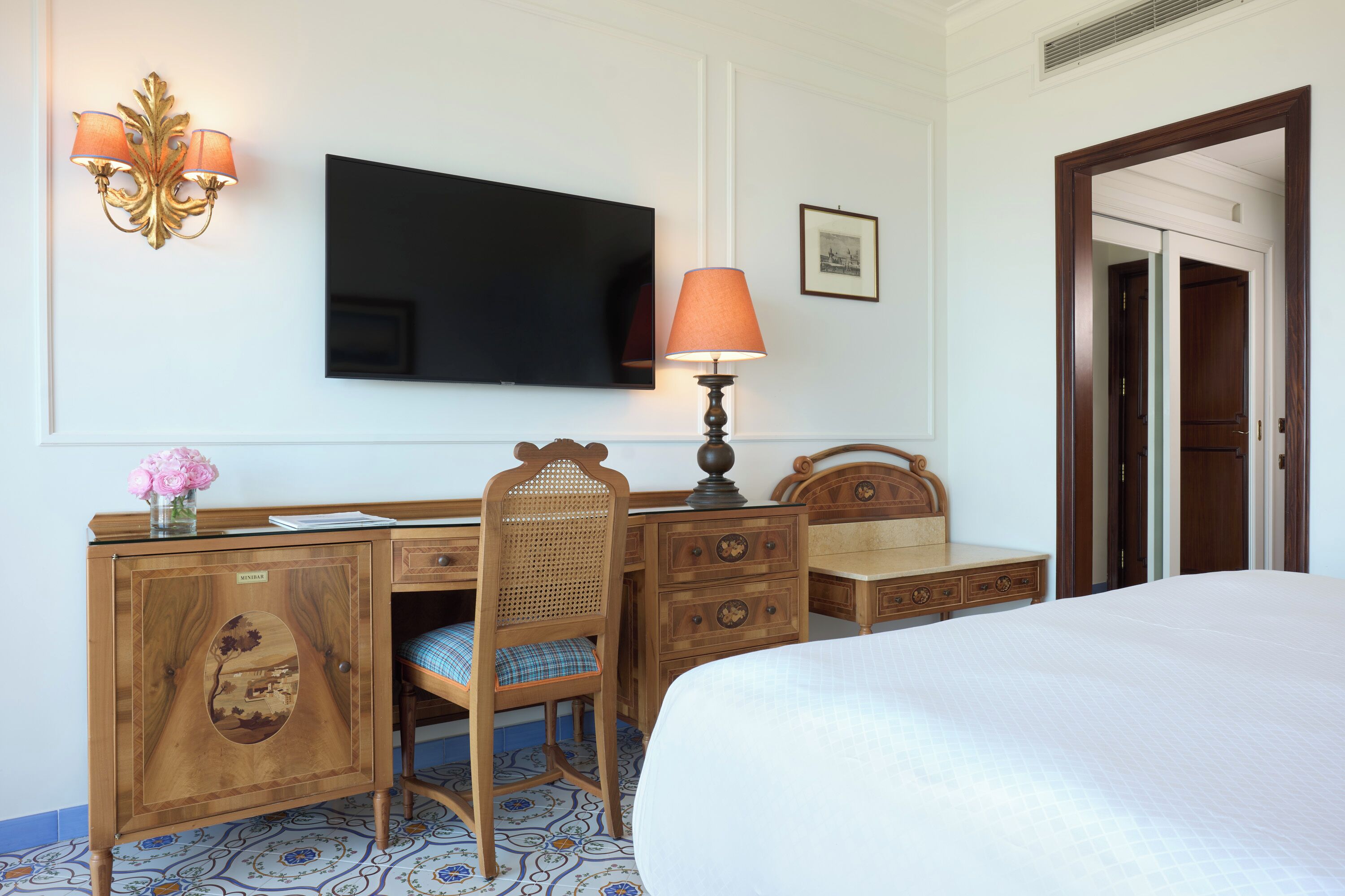 A hotel room with a bed, a desk with a chair, a television, a lamp, and a door.