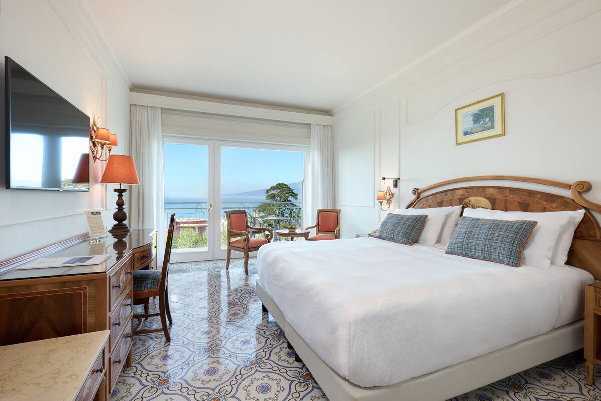 A hotel room with a large bed, a desk with a chair, a TV, and a balcony with a view of the sea.