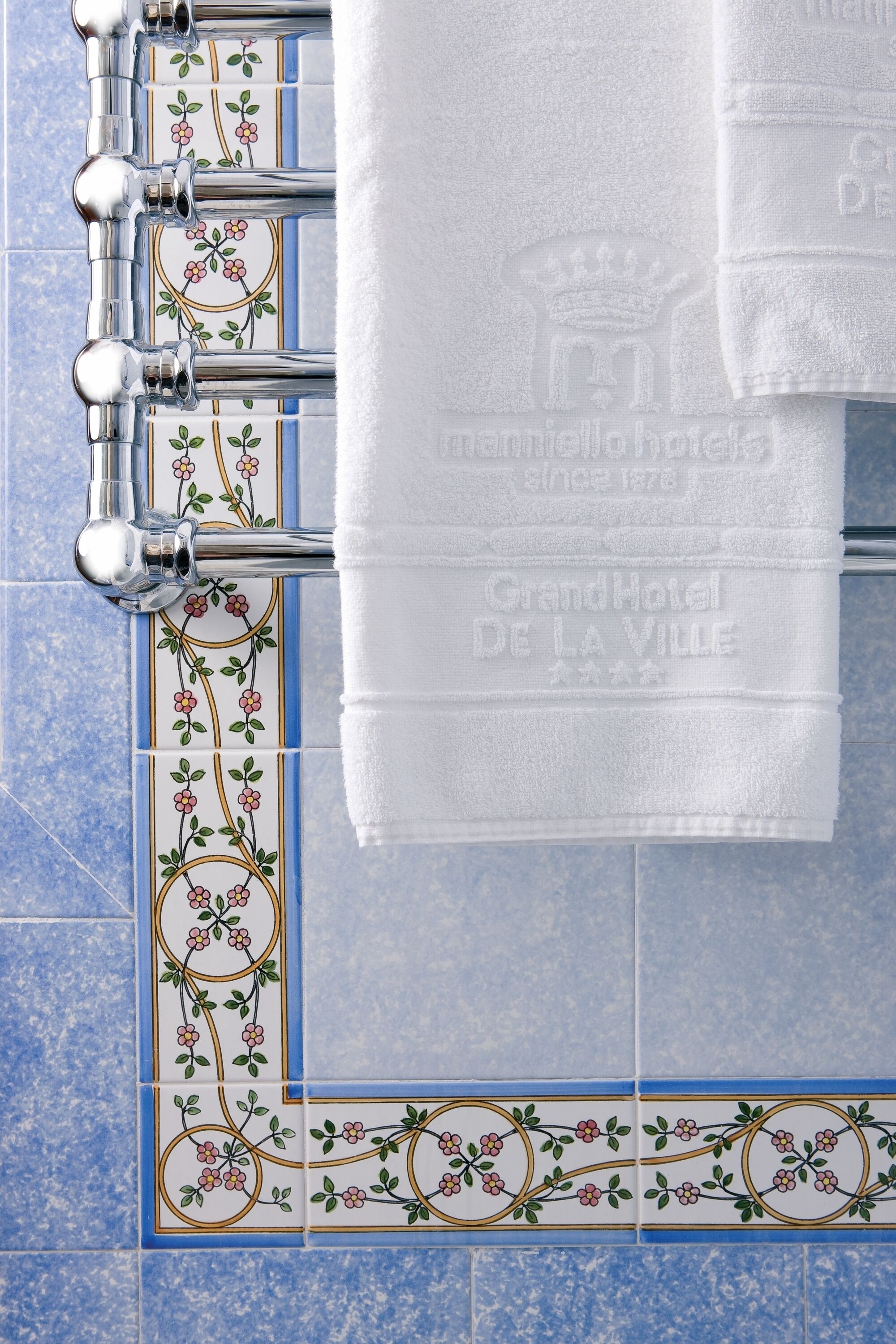 A bathroom with a towel rack holding two white towels, a decorative tile border, and a blue tiled wall.