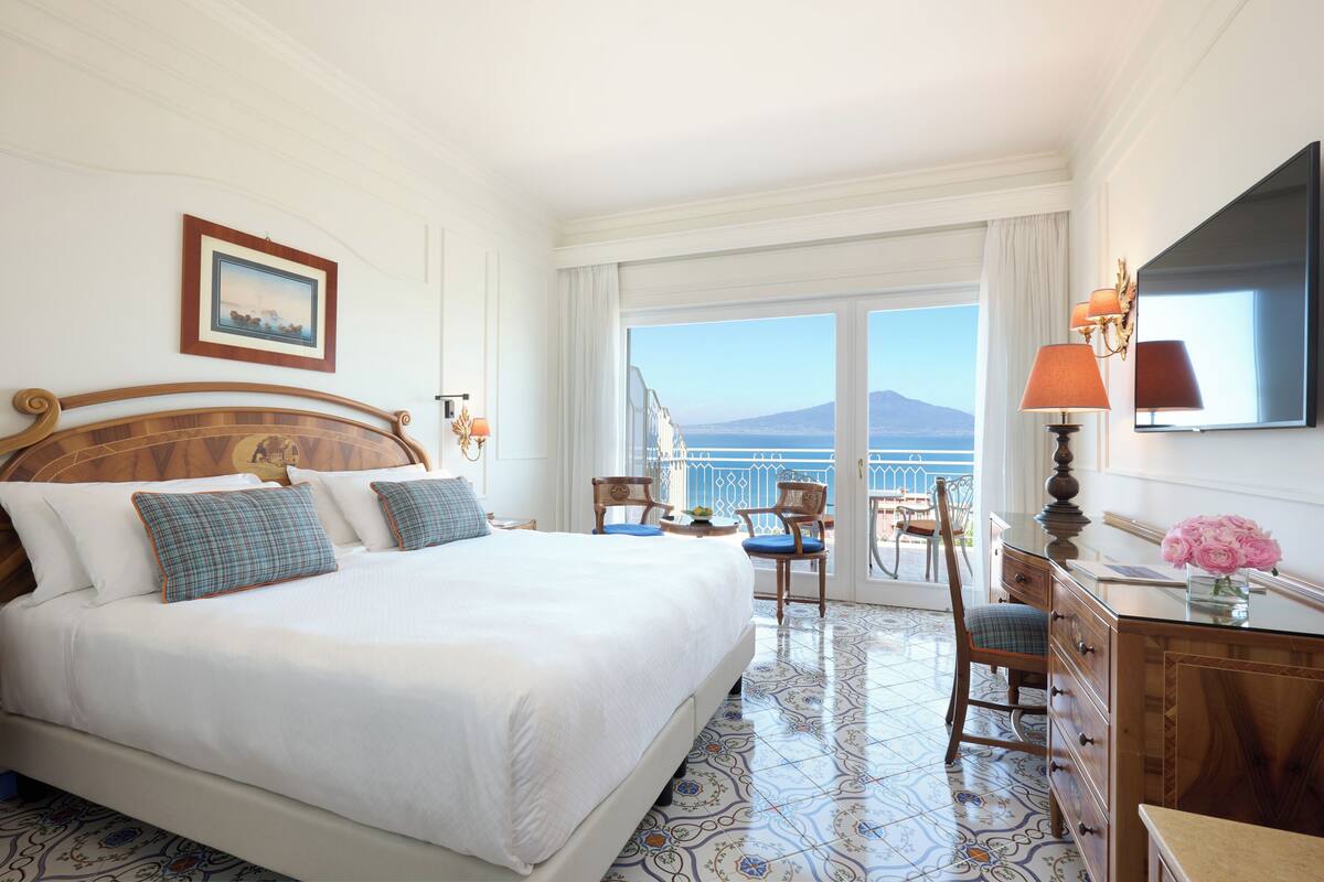 A hotel room with a large bed, a desk with a lamp, a chair, a TV, and a view of the sea.