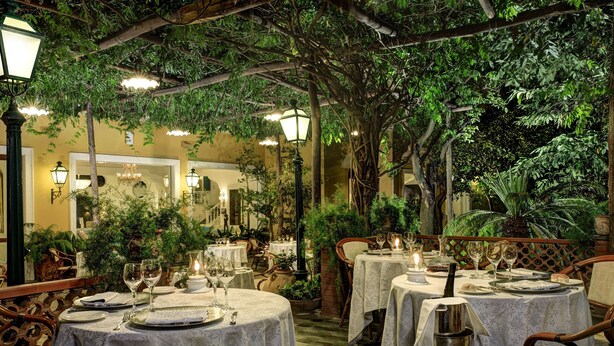Un área de comedor al aire libre con mesas preparadas para dos personas, rodeada de abundante vegetación e iluminación cálida.