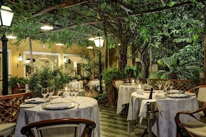 An outdoor dining area with tables set for two, surrounded by lush greenery and warm lighting. - Grand Hotel De La Ville (Sorrento)