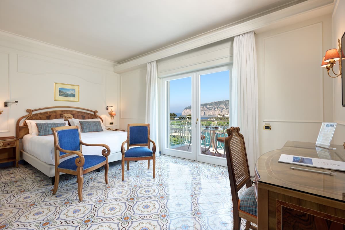 A hotel room with a bed, two chairs, a desk, and a large window with a view of the sea.
