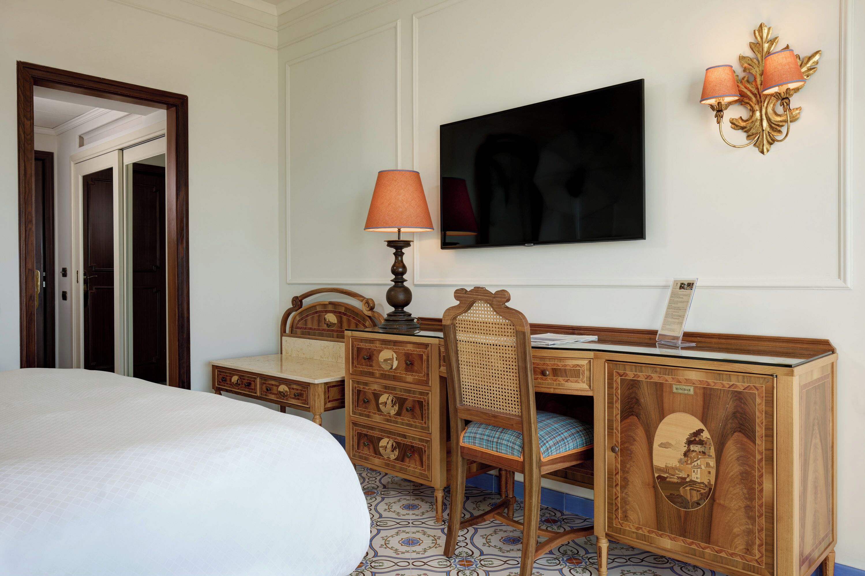 A hotel room with a bed, wooden desk, chair, television, lamp, and wall sconces.