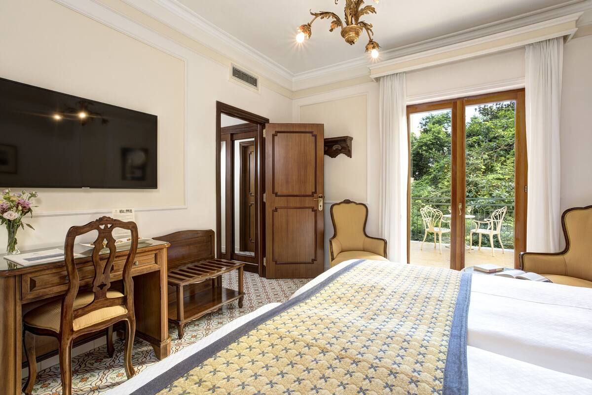 A hotel room with a large bed, a desk with a chair, a TV, and a balcony with chairs.