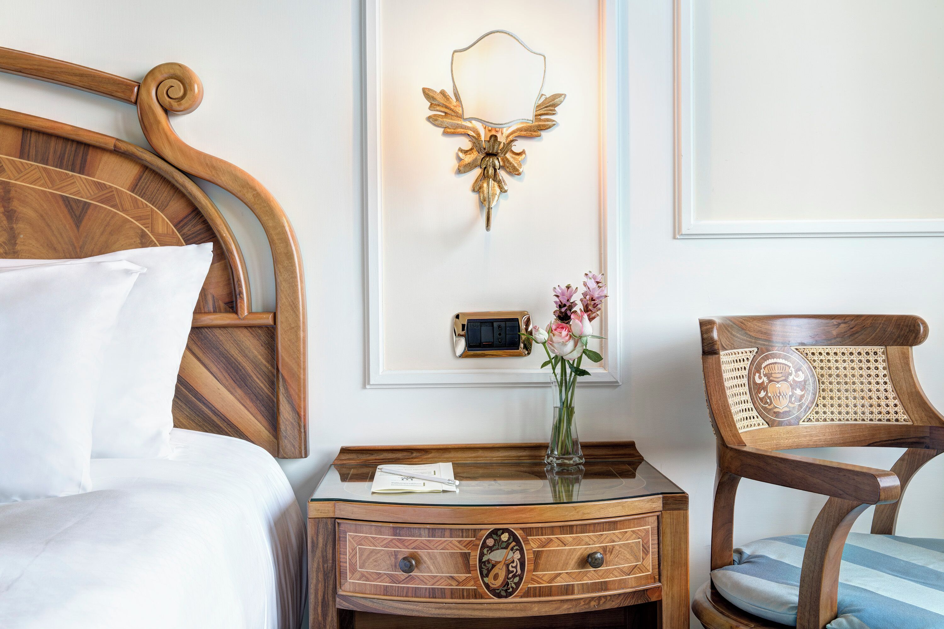 Un lit bien fait, avec du linge de lit blanc, une table de chevet en bois sur laquelle se trouvent une radio et un vase de fleurs, et une chaise en bois avec un coussin.