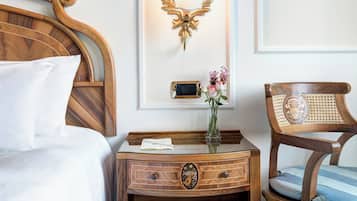 Ein ordentlich bezogenes Bett mit weißen Leinen, ein Nachttisch aus Holz mit einem Radio und einer Blumenvase sowie ein Holzstuhl mit Kissen.