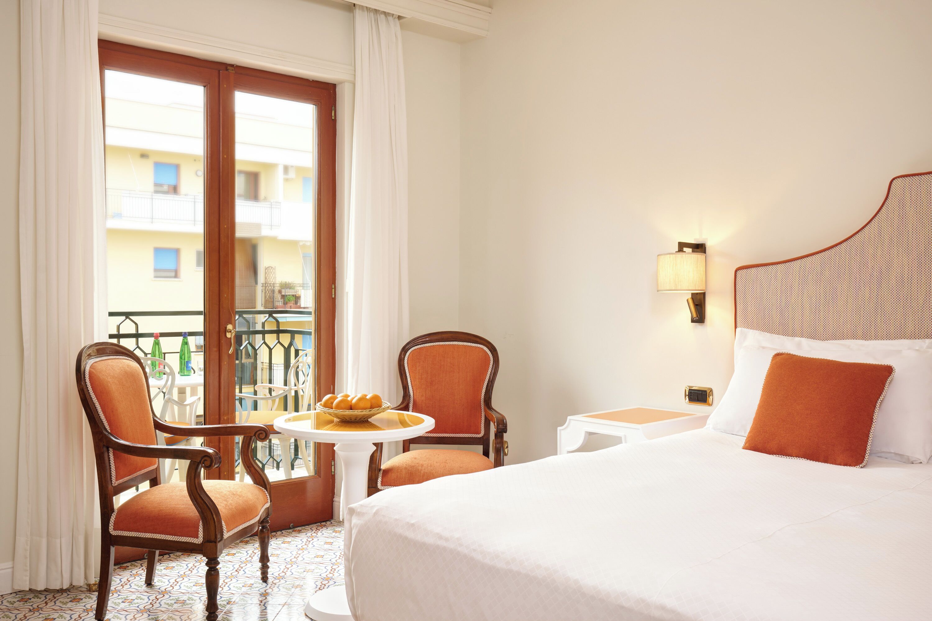 A hotel room with a bed, two chairs, a small table with a fruit bowl, a nightstand, and a view of a balcony with a railing.
