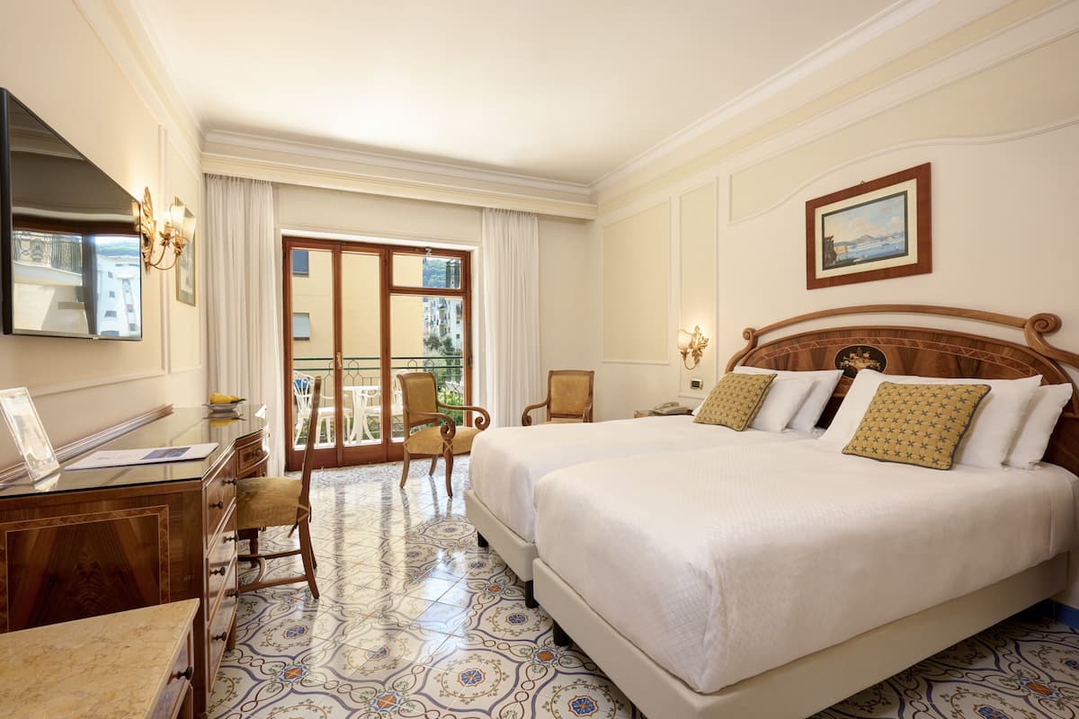 A hotel room with a large bed, a desk, a chair, a balcony with a view of buildings, and a painting on the wall.