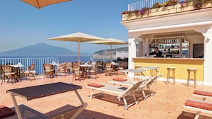 Una terraza con sillones de descanso, sombrillas y una zona de bar con vistas al mar y a la montaña.