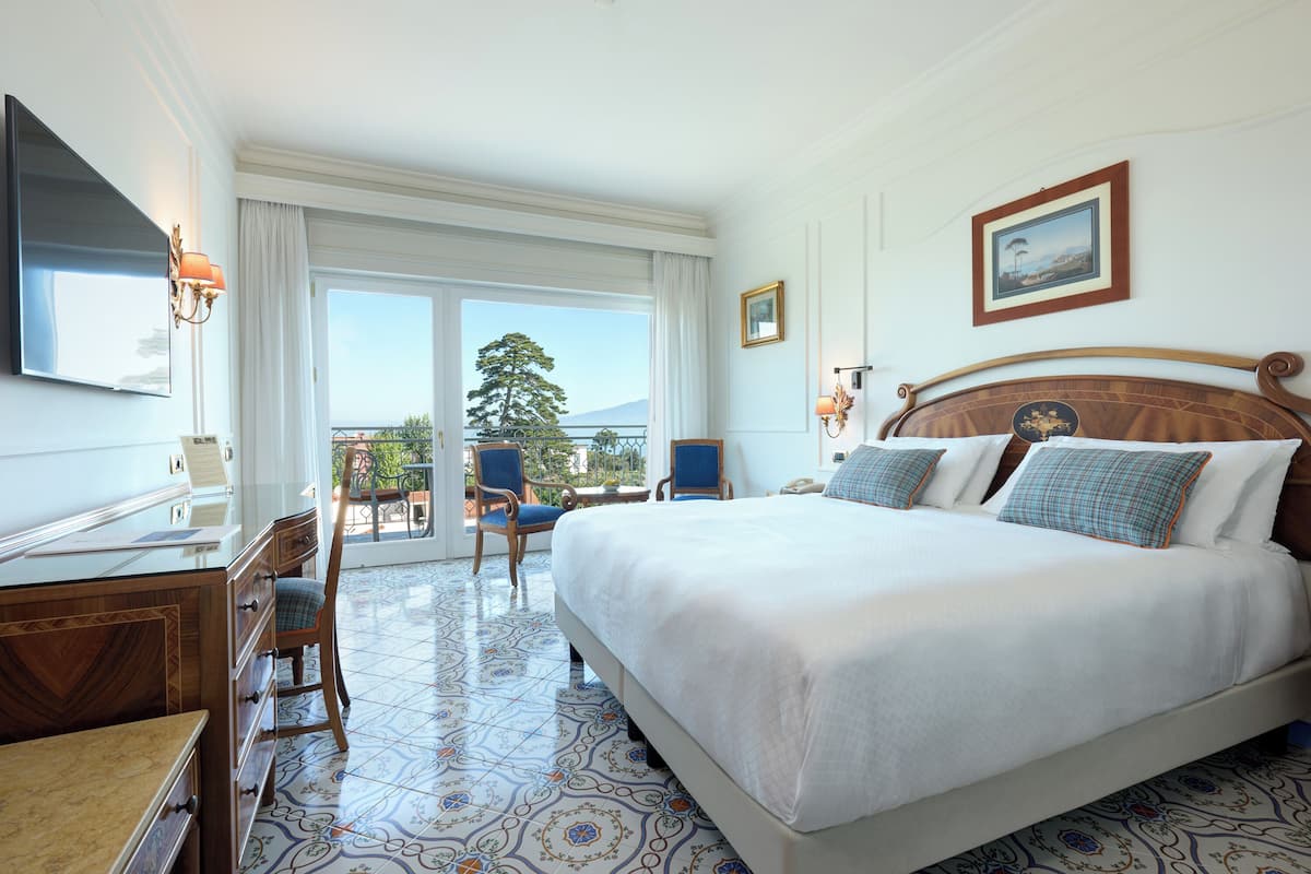 A hotel room with a large bed, a desk with a chair, a TV, and a balcony with a view of the outdoors.
