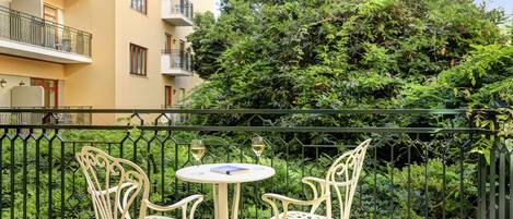 A balcony with two white metal chairs and a small round table, surrounded by a green metal fence and dense foliage.