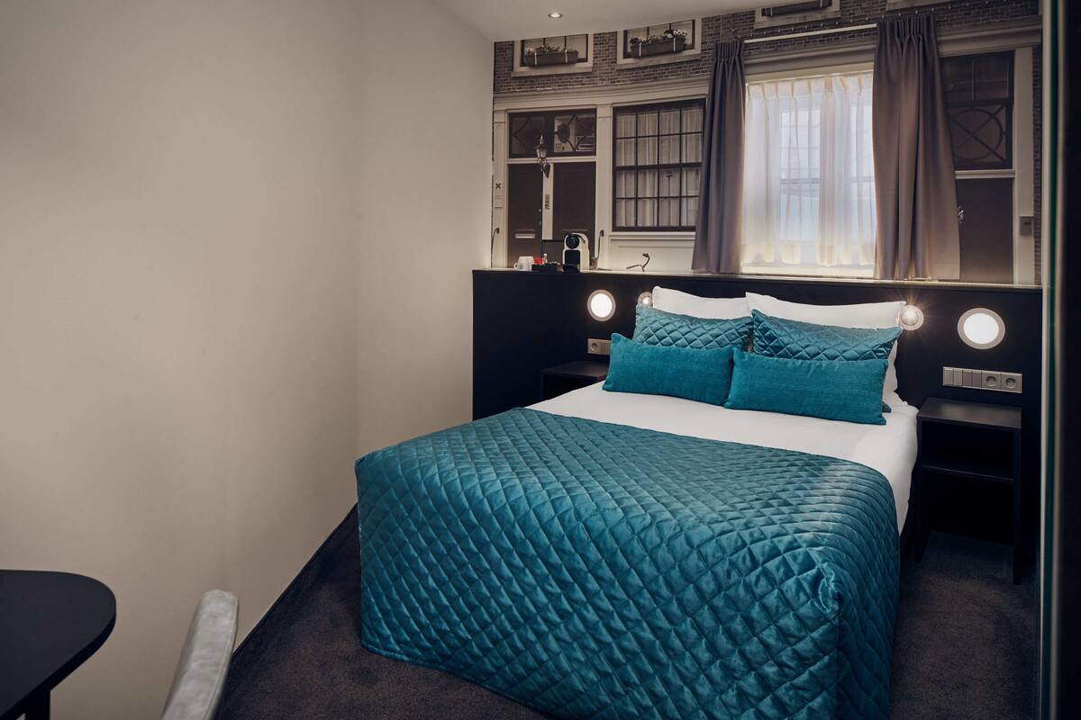 A hotel room with a bed, bedside tables, a nightstand, a window with curtains, and a wall-mounted shelf with decorative items.