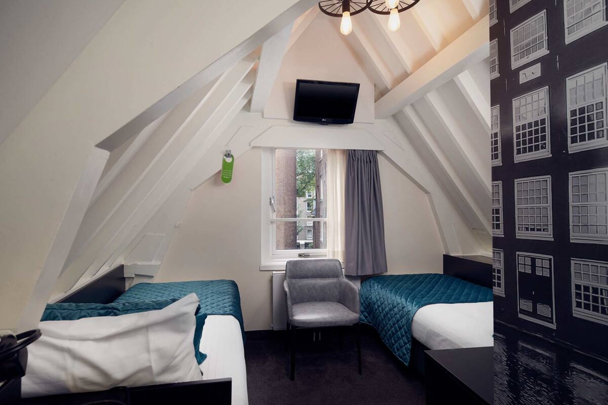 A dormitory room with two beds, a chair, a TV, and a window with a view of buildings.