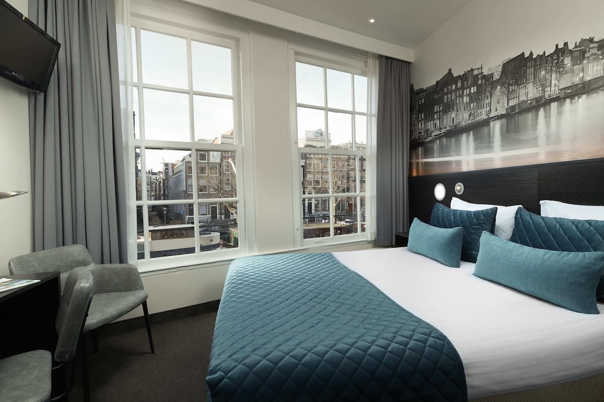 A hotel room with a large bed, a desk, and a chair. There are two windows with a city view.