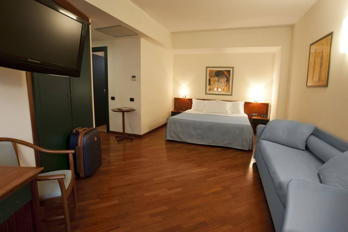 A hotel room with a bed, a sofa, a desk with a chair, a television, and a painting on the wall.