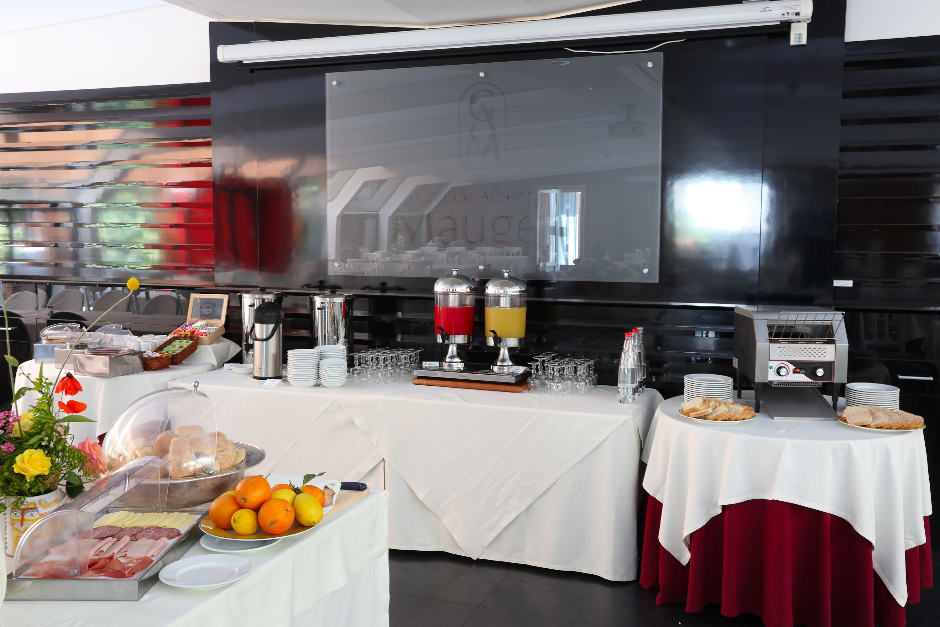 Montaje de bufé con diversas opciones de comida, una estación de café y una pantalla grande que muestra el nombre del hotel.