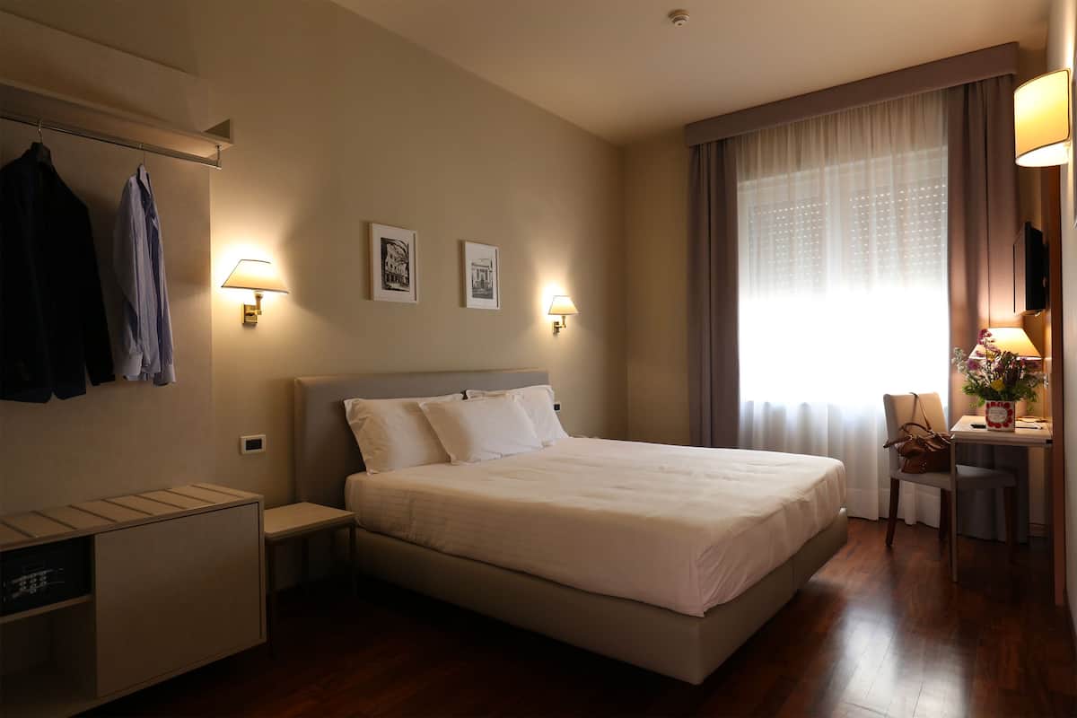 A hotel room with a large bed, bedside lamps, a desk with a chair, and a window with curtains.