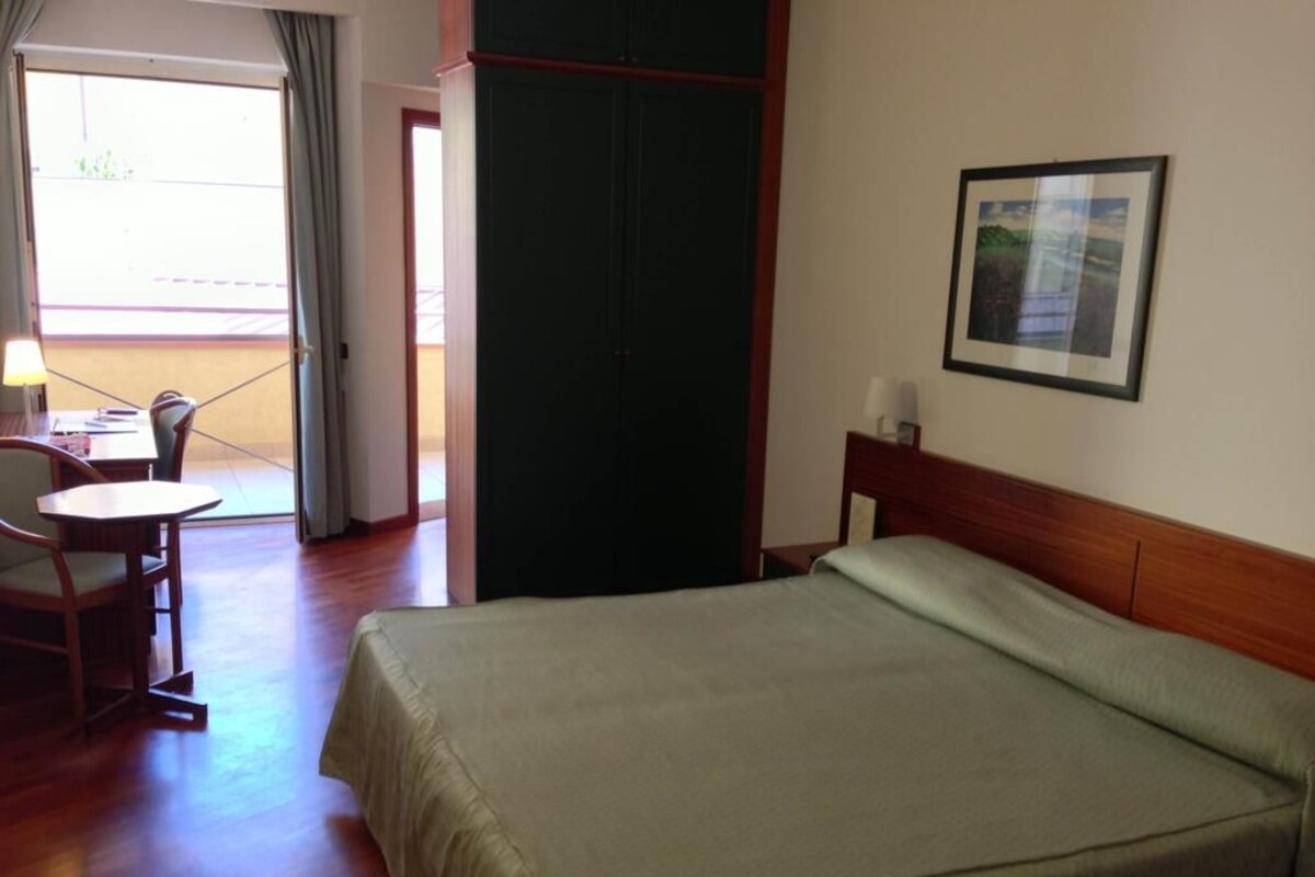 A hotel room with a bed, a desk, and a chair. There is a window with curtains, a door, and a framed picture on the wall.