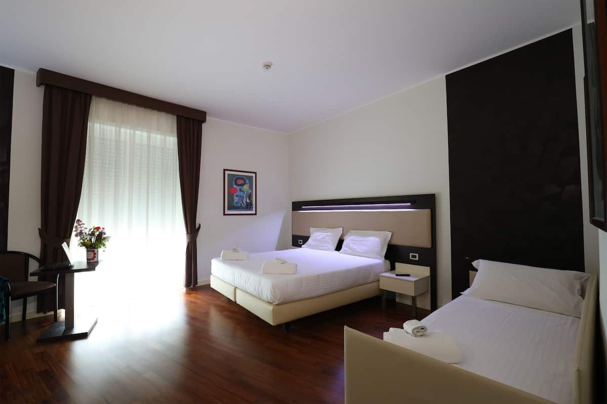 A modern hotel room with a large bed, a desk with a laptop, a chair, and a bedside table with a lamp.