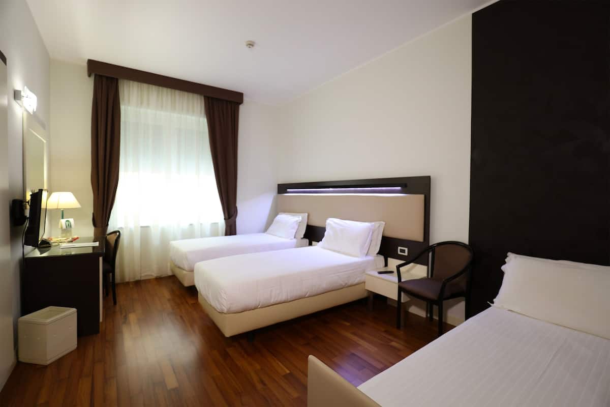 A hotel room with two beds, a desk with a lamp, a chair, and a large window with curtains.