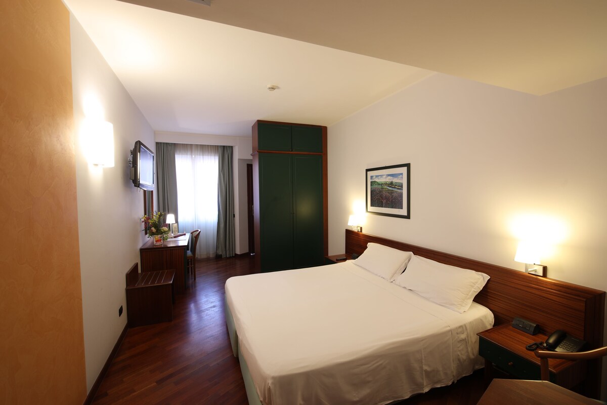 A hotel room with a bed, a desk with a chair, a wardrobe, and a painting on the wall.