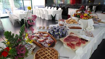 Montaje de bufé con diferentes opciones de comida, incluyendo pasteles, carnes y frutas, dispuestas sobre mesas cubiertas con manteles blancos.