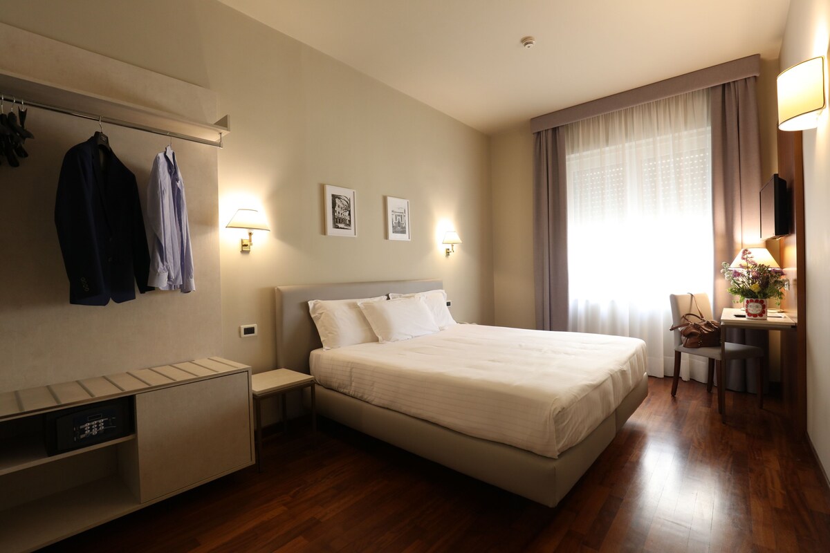 A hotel room with a bed, a bedside table, a lamp, a chair, a desk, a mirror, and a wardrobe.