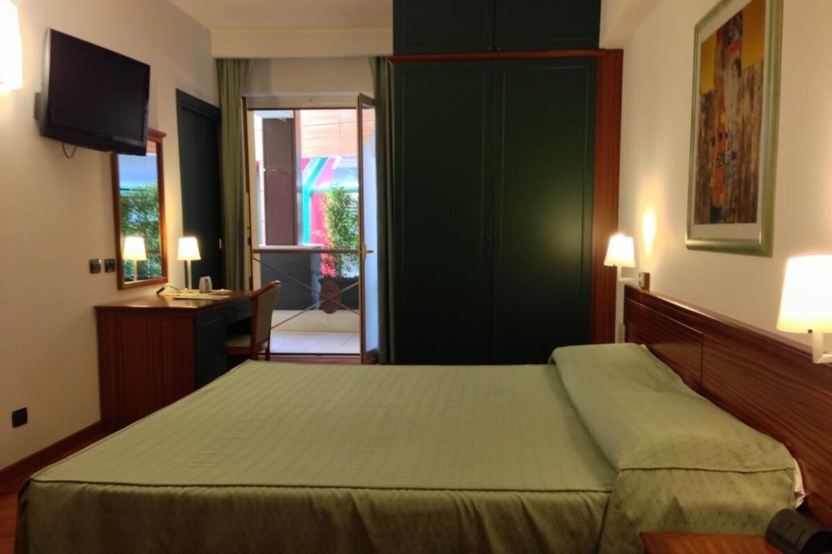 A hotel room with a bed, bedside lamps, a desk with a telephone, a television on the wall, and a door leading to an outdoor area.