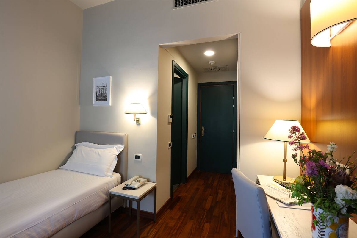A hotel room with a bed, bedside table, lamp, chair, and a desk with a flower vase.