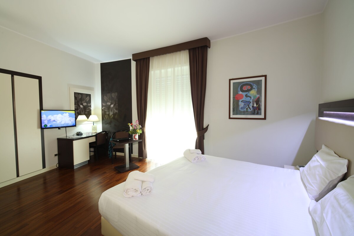 A hotel room with a bed, a desk, a chair, a television, and a painting on the wall.