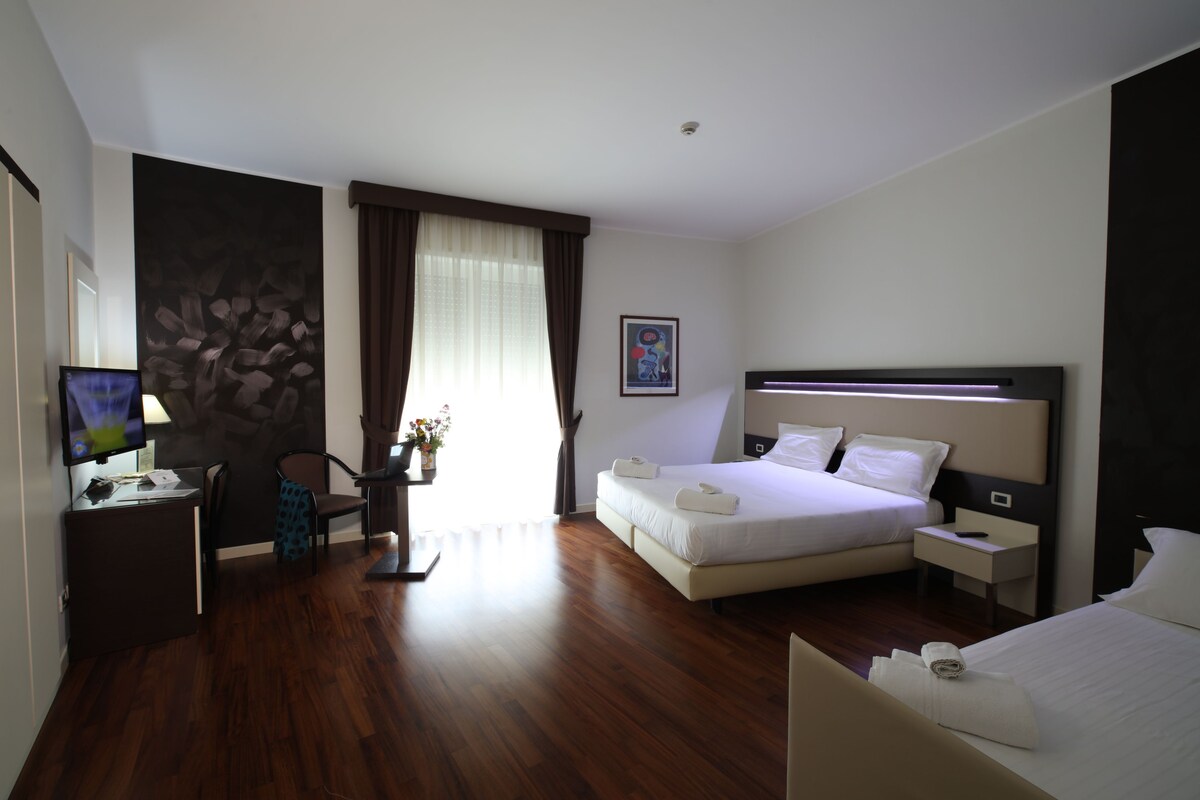 A hotel room with a bed, bedside tables, a desk with a chair, a television, and a painting on the wall.