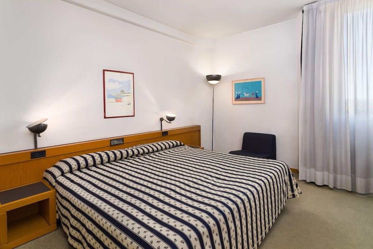 Une chambre d’hôtel comprenant un lit, un bureau, une chaise, deux lampes fixées au mur et deux tableaux encadrés.