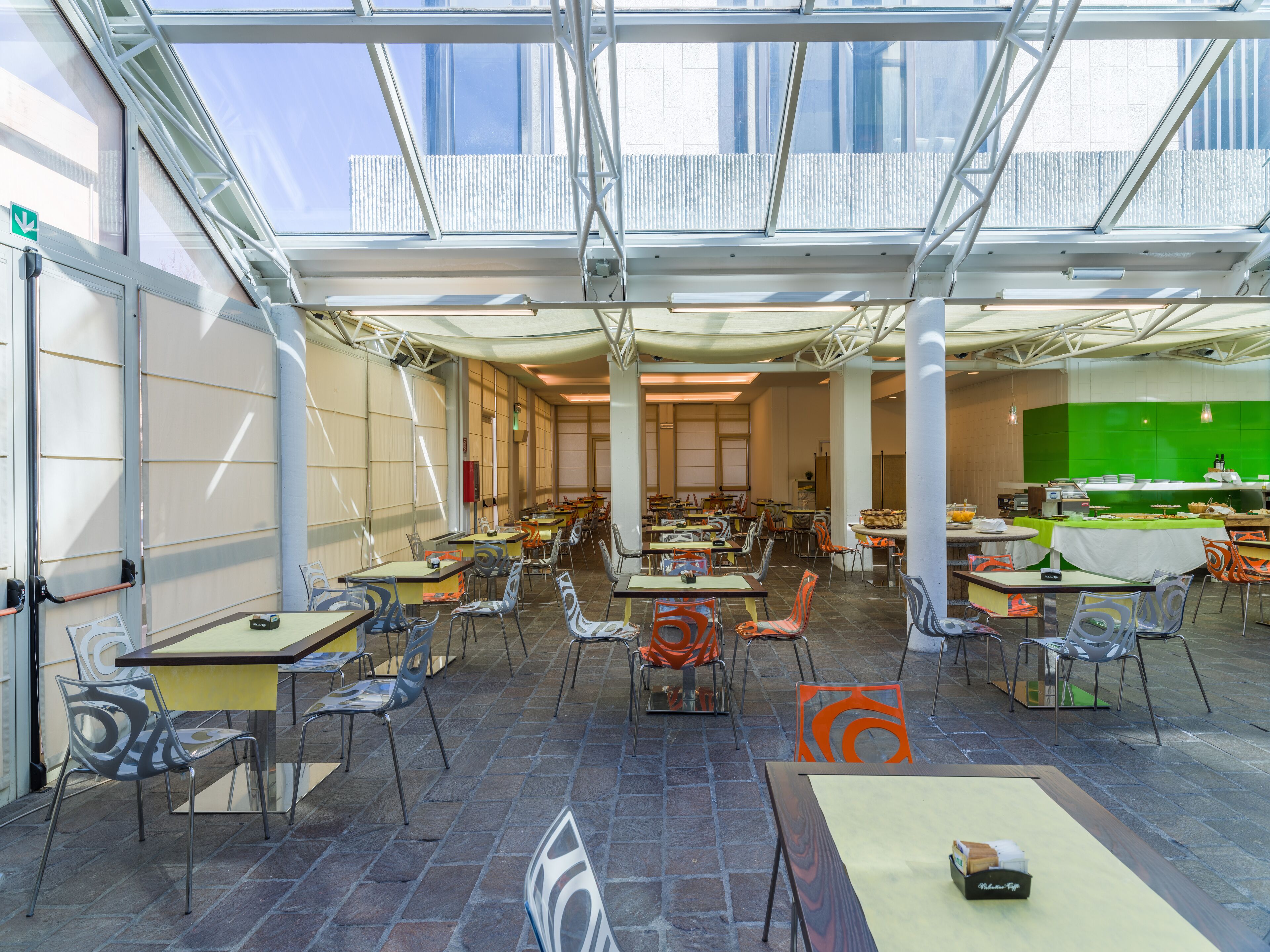 A spacious indoor dining area with high ceilings and large windows, featuring multiple tables and chairs arranged for dining.