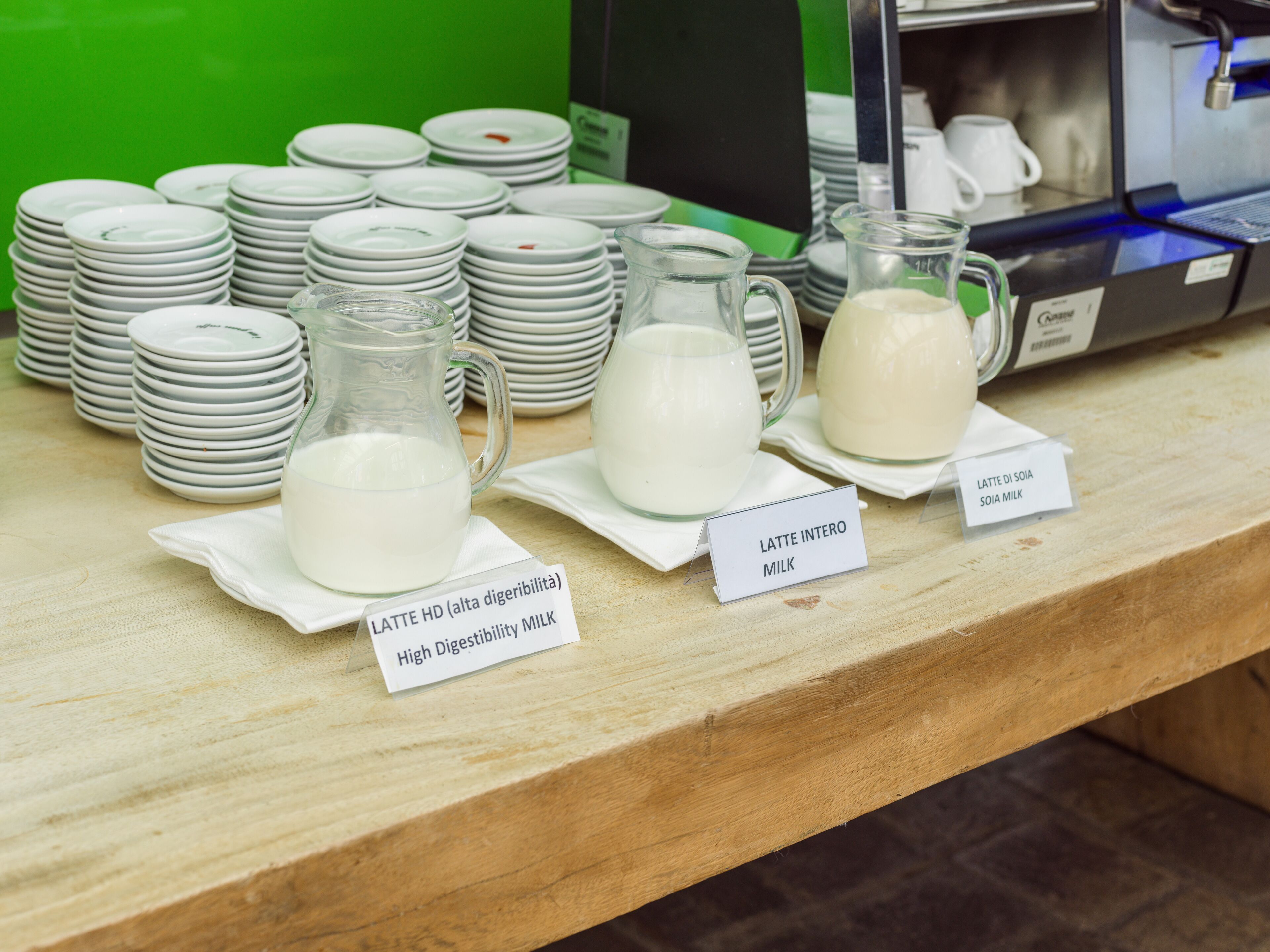 Een koffiehoek met stapels witte borden, voorzien van labels met verschillende soorten melk, en een koffieapparaat op de achtergrond.