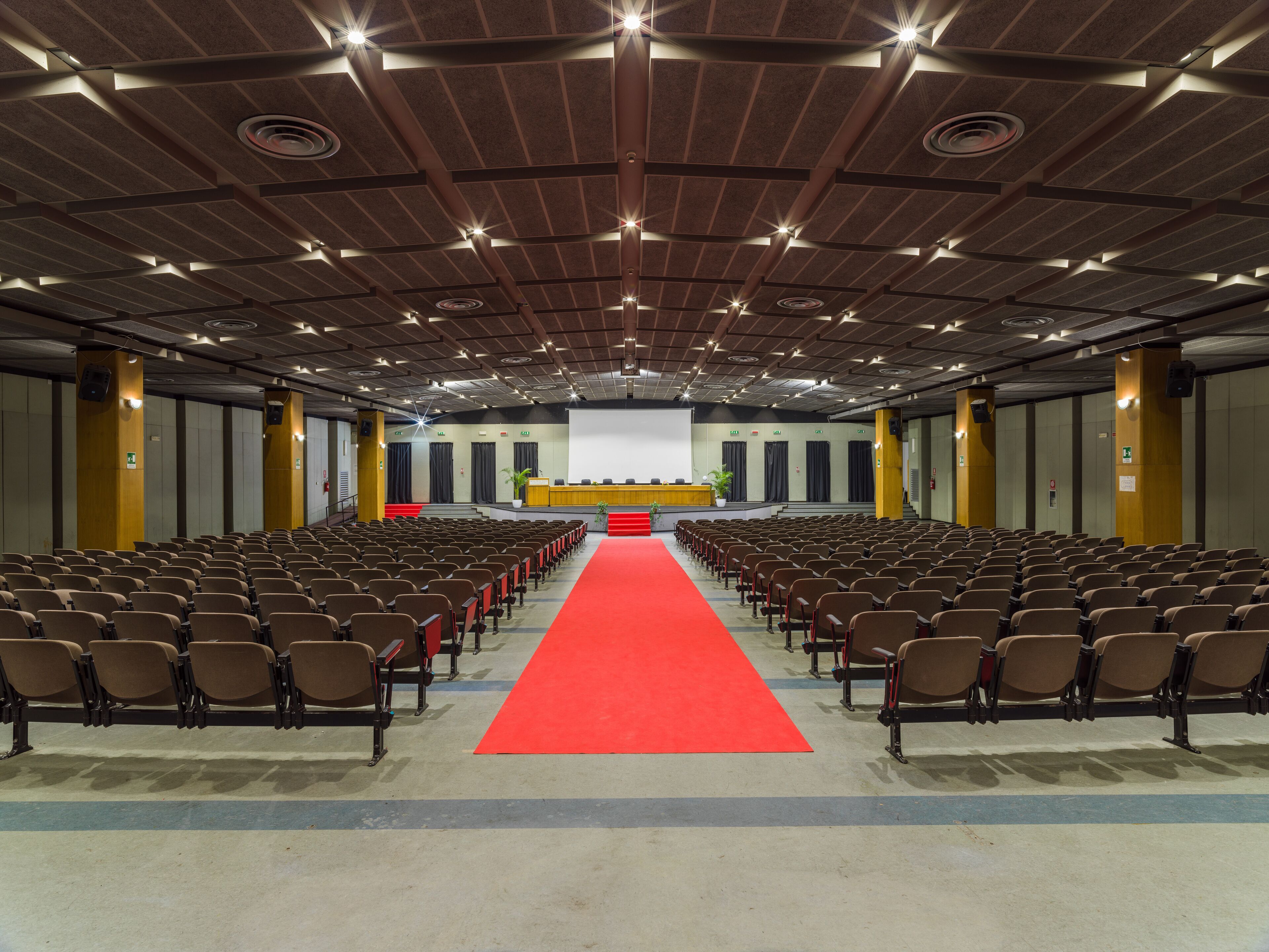 A large conference room with rows of chairs, a red carpet, and a stage with a screen at the front.