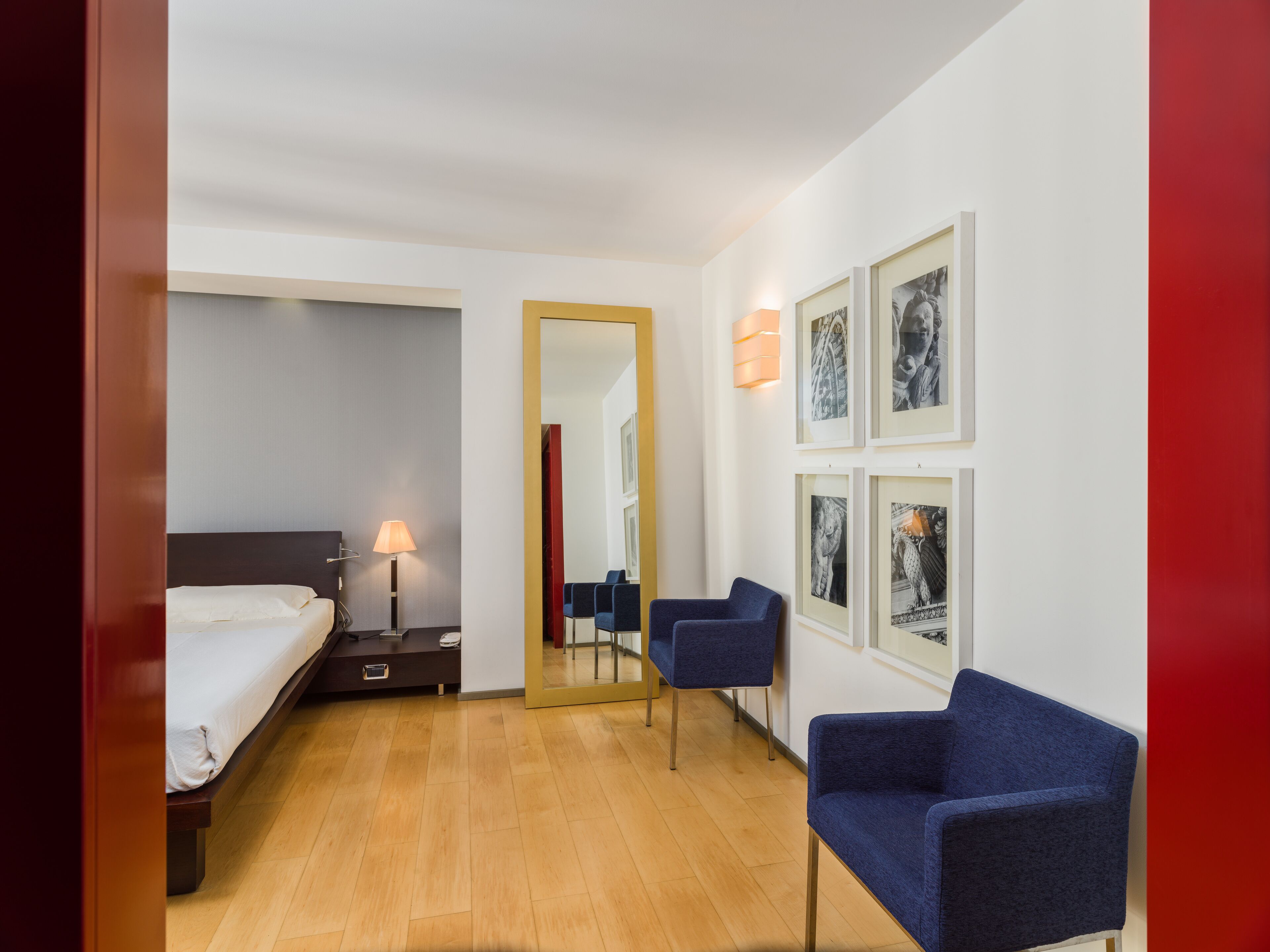 A hotel room with a bed, bedside table, lamp, two chairs, and framed pictures on the wall.