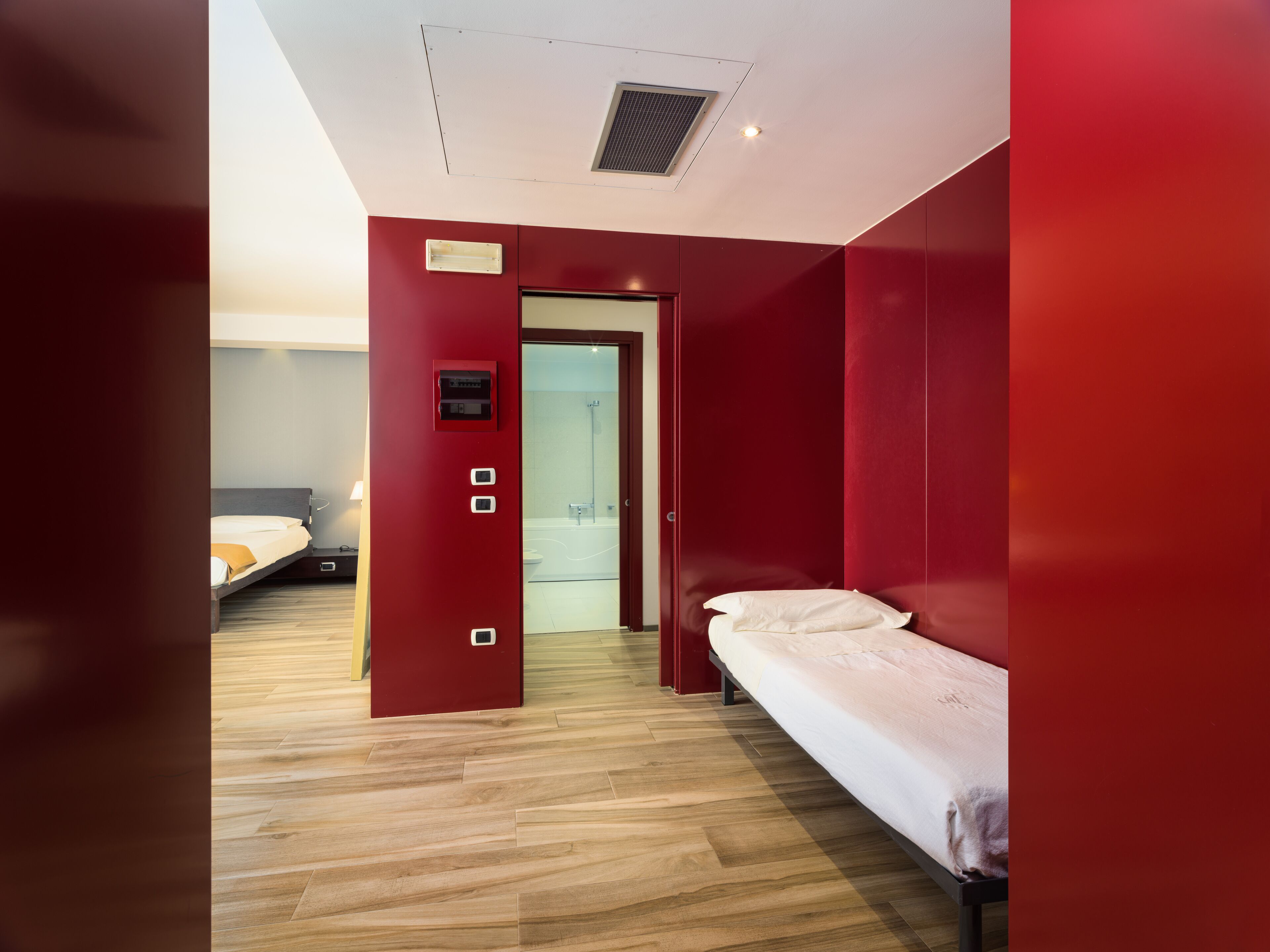 Habitación de hotel con piso de madera, cama con ropa de cama blanca, pared de acento roja y una mesita de noche pequeña.