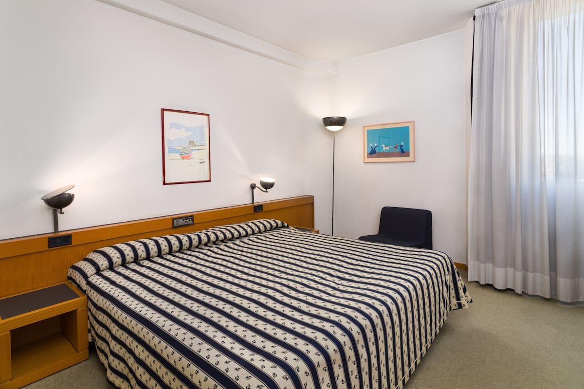 Habitación de hotel con cama, una silla, un escritorio y dos lámparas fijadas a la pared.