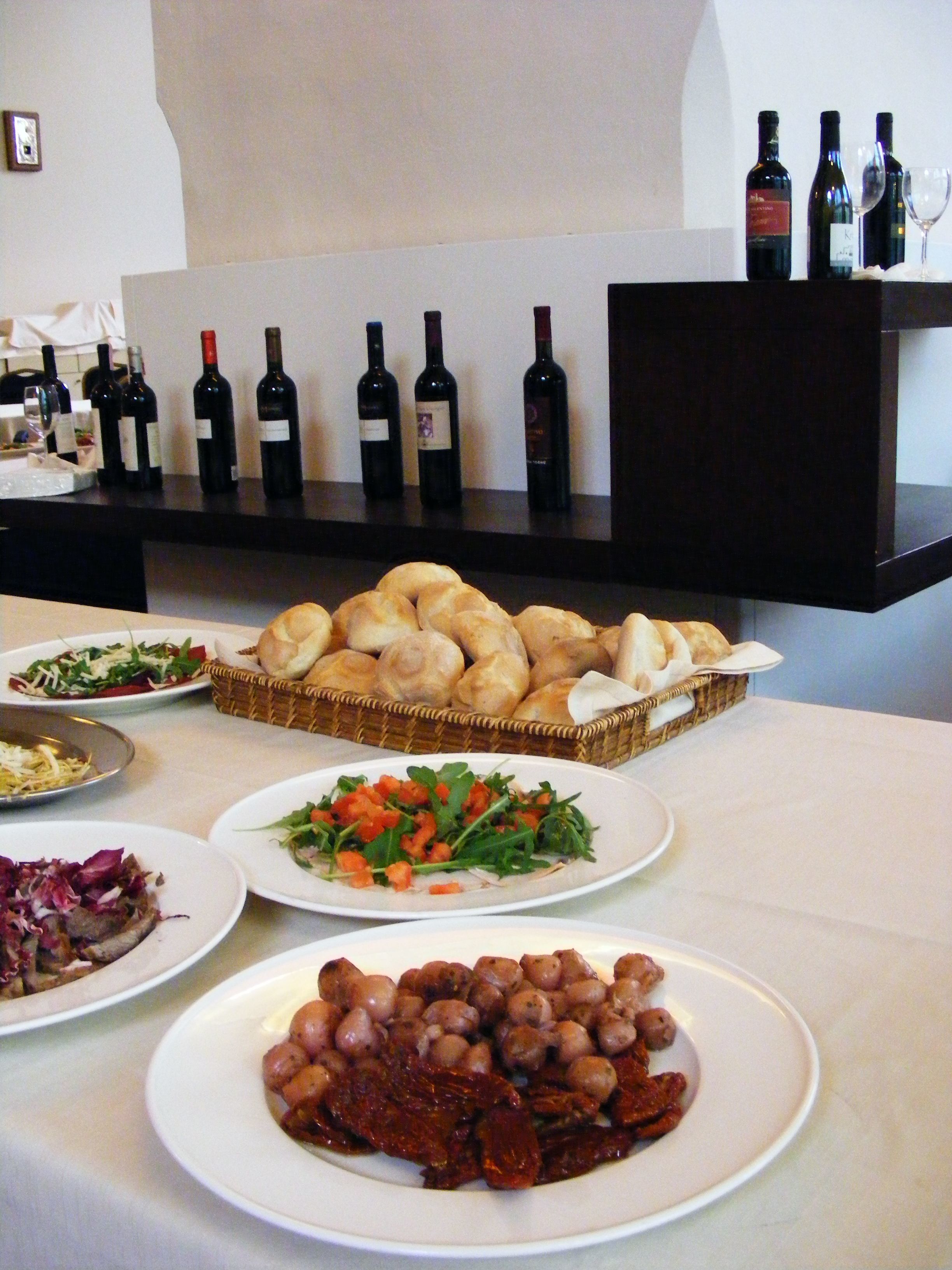Una mesa de buffet con diversos platos, entre ellos ensaladas, carnes y pastelería, y una selección de botellas de vino al fondo.