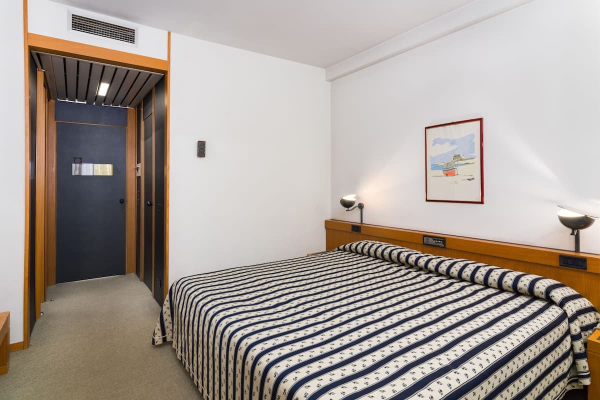 Una habitación de hotel con una cama, una puerta, una lámpara de pared y un cuadro enmarcado.