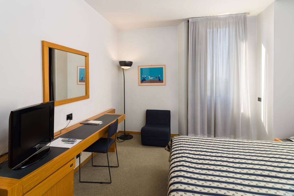 Una habitación de hotel con una cama, un escritorio con un ordenador, una silla, un televisor, una lámpara y un cuadro en la pared.