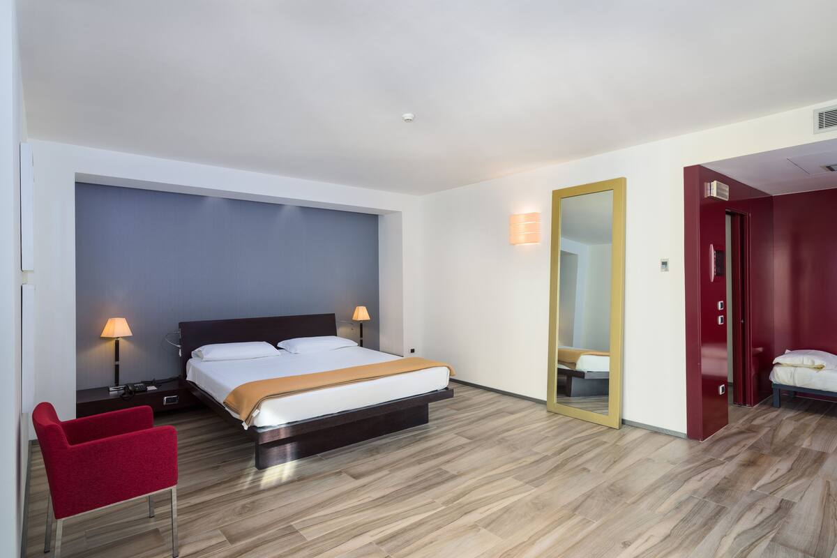 Una habitación de hotel moderna con una cama grande, lámparas de noche, una silla roja y un espejo en la pared.