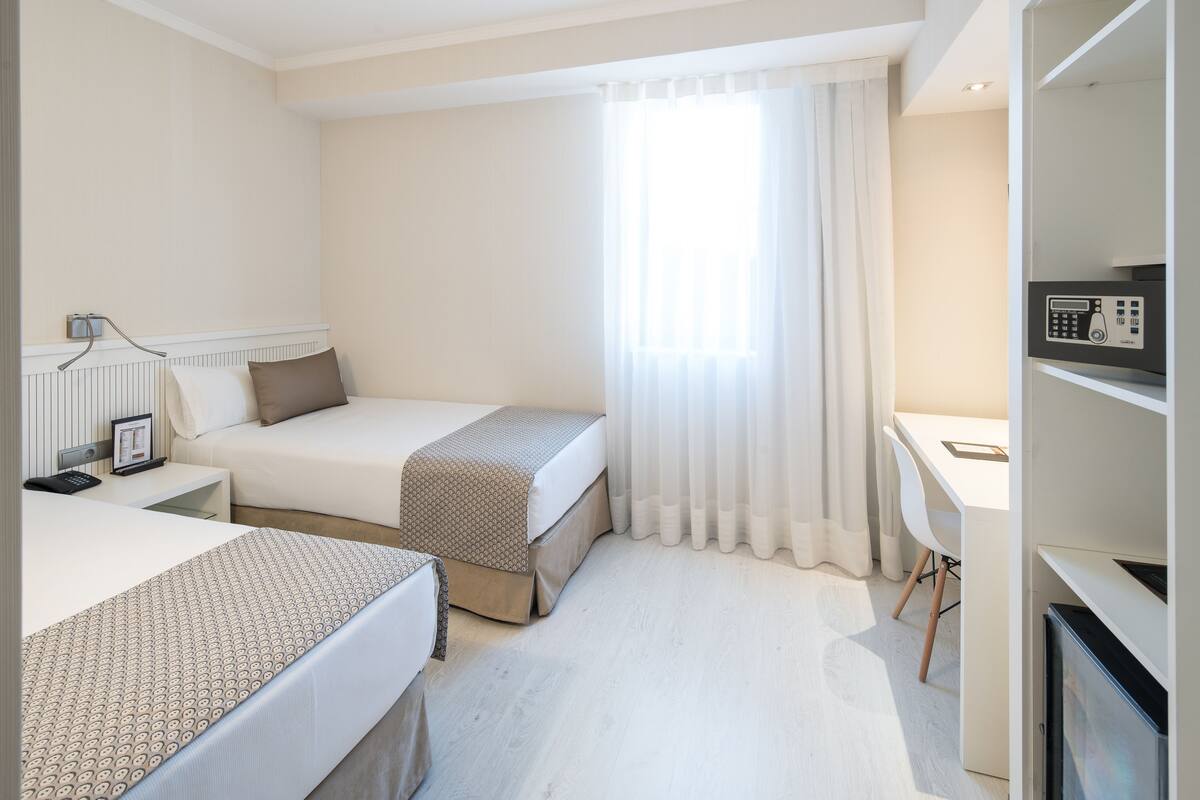 Ein Hotelzimmer mit zwei Betten, einem Schreibtisch, einem Stuhl, einem Fernseher und einem Fenster mit Vorhängen.