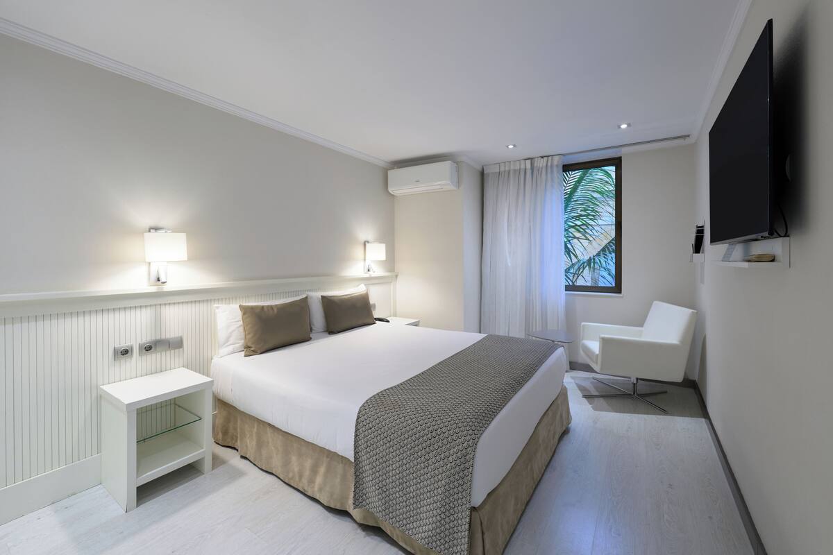 Ein modernes Hotelzimmer mit einem großen Bett, Nachttischlampen, einem Flachbildfernseher, einem Fenster mit Vorhängen und einem weißen Stuhl.
