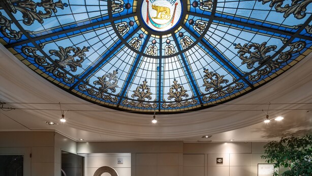 Un comptoir d’accueil avec un plateau en verre, un grand miroir décoratif de forme circulaire au-dessus, et un plafond en vitrail bleu orné d’un emblème représentant un lion.