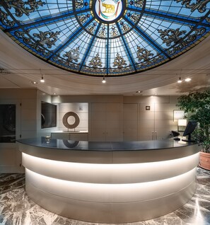 A reception desk with a glass top, a circular decorative mirror above it, and a blue stained glass ceiling with a lion emblem. - Catalonia Roma (Barcelona)