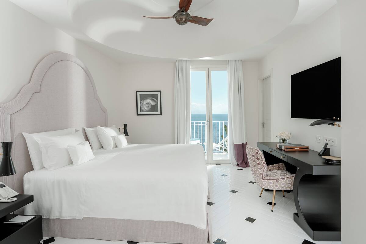 A modern hotel room with a large bed, a desk with a chair, a ceiling fan, and a view of the ocean through the balcony doors.